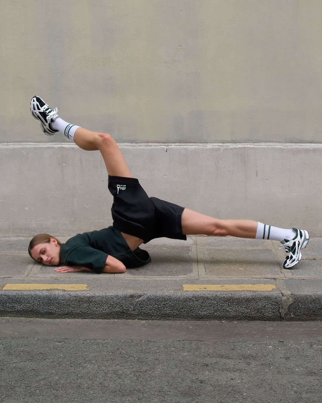 A woman in a dark green t-shirt and black shorts lying on pavement with legs raised, photographed by Yis Kid in Berlin.