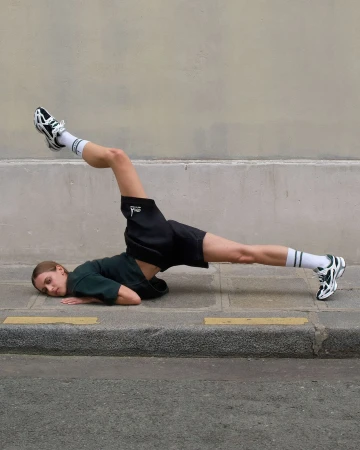 A woman in a dark green t-shirt and black shorts lying on pavement with legs raised, photographed by Yis Kid in Berlin.