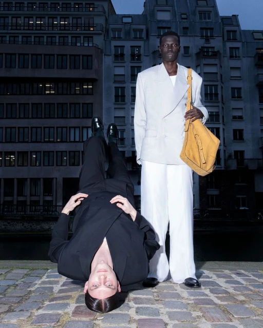 Two people pose against a city building. One stands holding a bag and wears white suit, the other is laying down in a black suit.