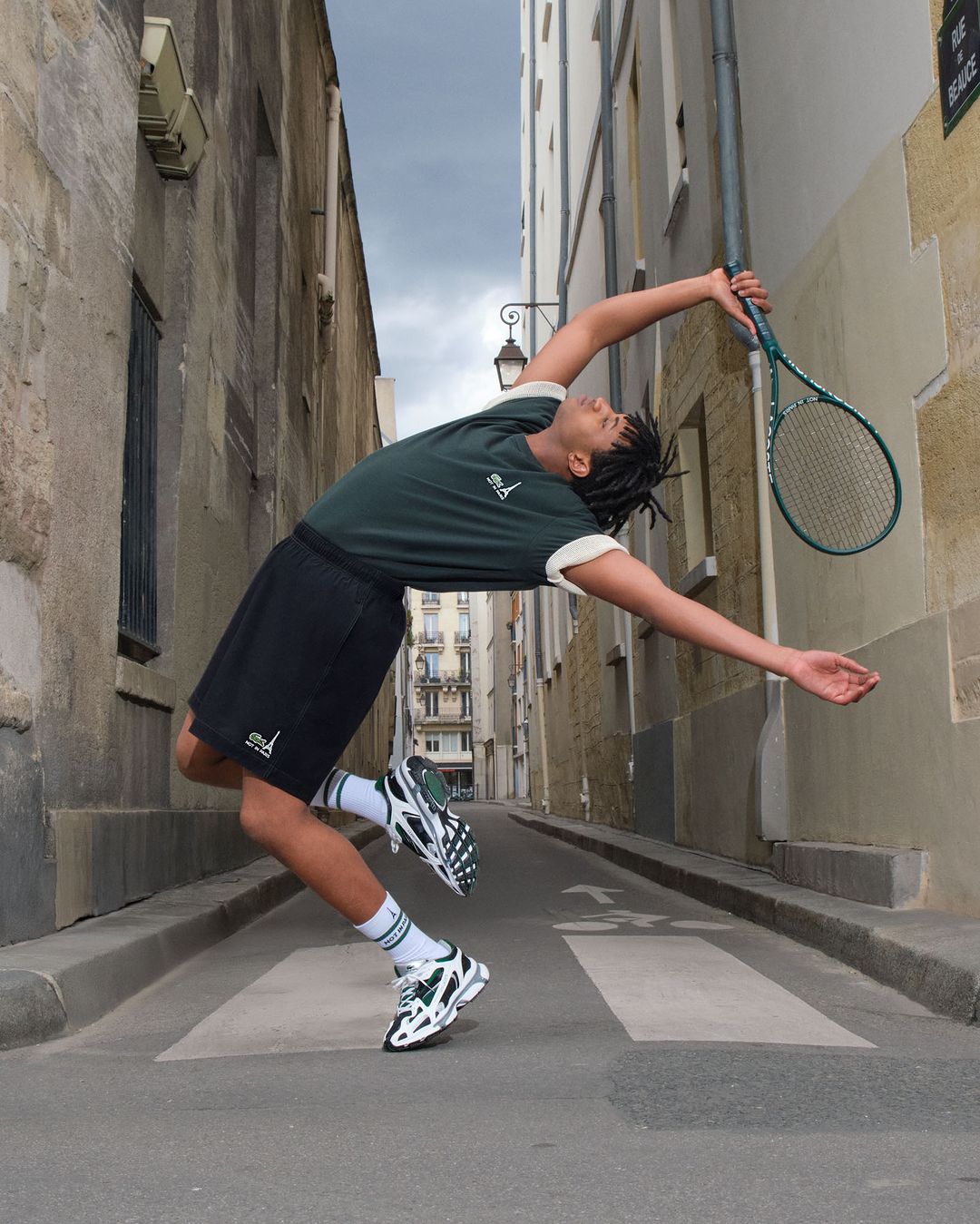 Adult male with dreadlocks, wearing a green top and black shorts, leaning back while holding a tennis racket on a city street.