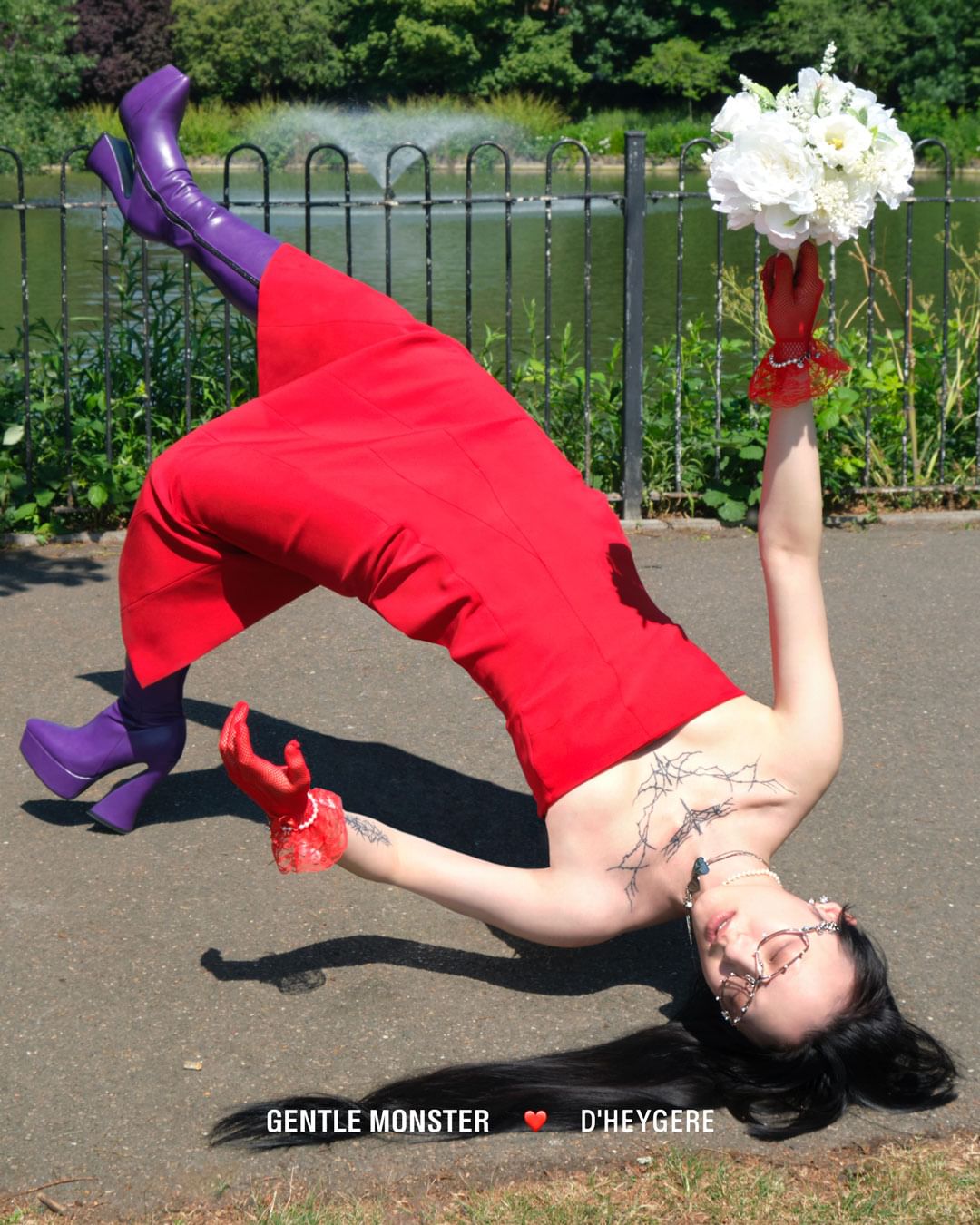 Woman in red dress and purple boots performs a backbend with a flower bouquet in a park setting on a sunny day.