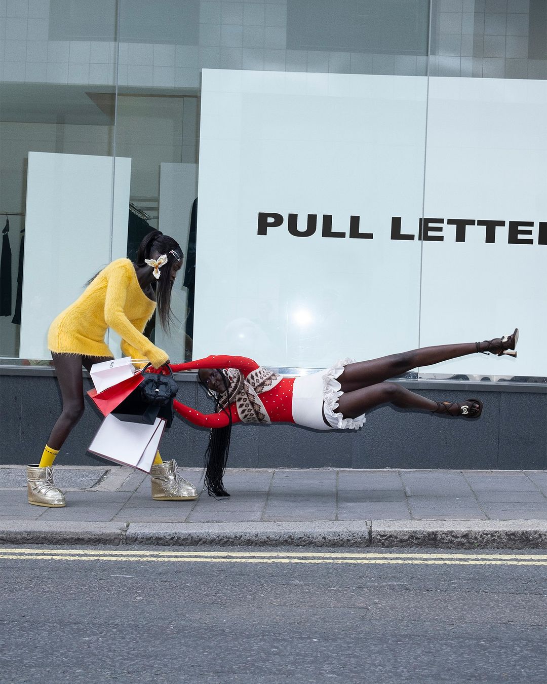 Two women stage a scene with one appearing to pull the other as she floats horizontally on a sidewalk in front of a building.
