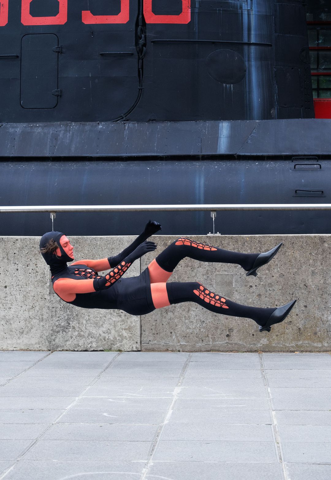 Person in black costume levitating outdoors in front of black building with red numbers, wearing gloves, thigh high socks, and heeled boots.