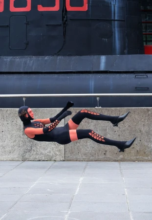 Person in black costume levitating outdoors in front of black building with red numbers, wearing gloves, thigh high socks, and heeled boots.