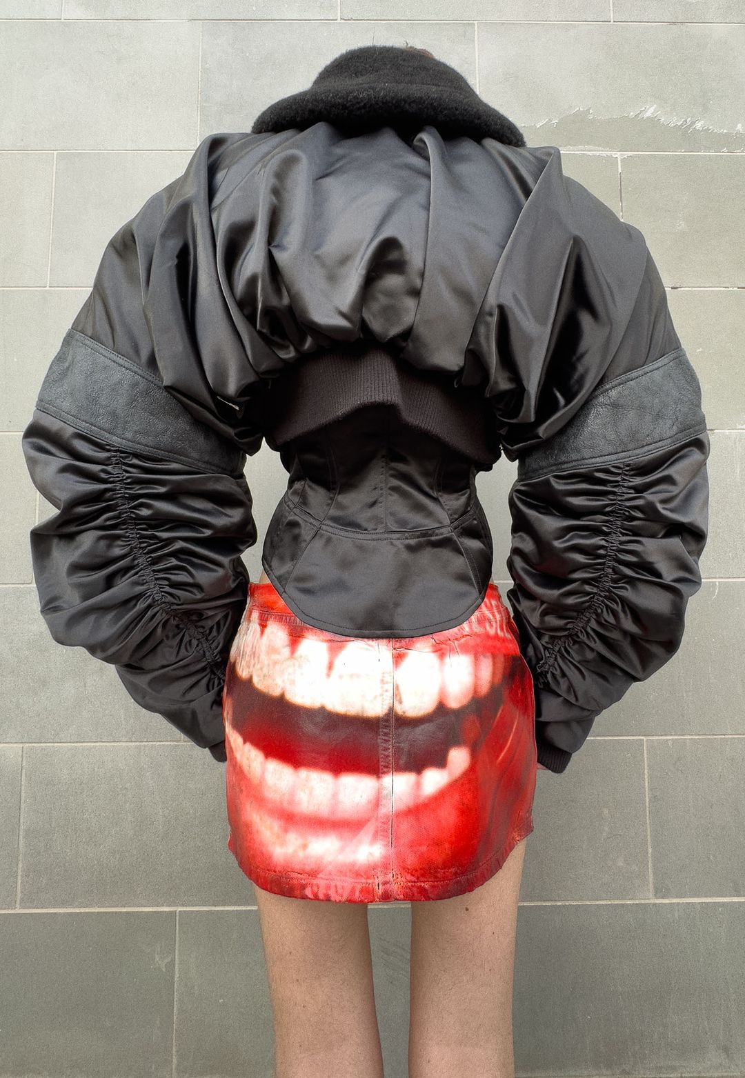 Back view of a person wearing a black bomber jacket with balloon sleeves and a mini skirt with the print of a large mouth.