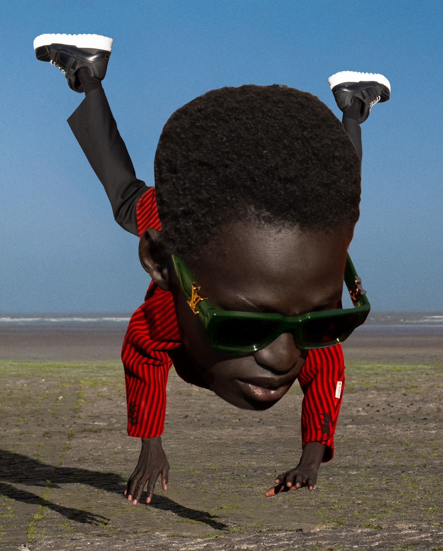An African American person in a red jacket, black pants, and sunglasses does a handstand on a beach with a blue sky and water background.