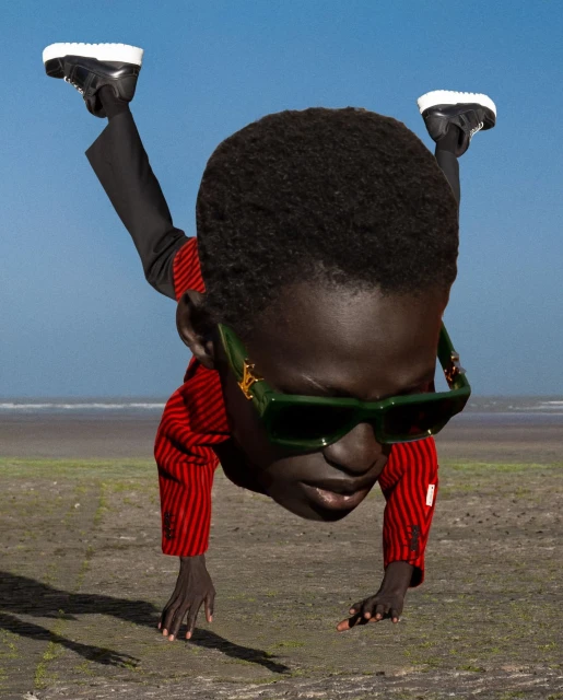 An African American person in a red jacket, black pants, and sunglasses does a handstand on a beach with a blue sky and water background.