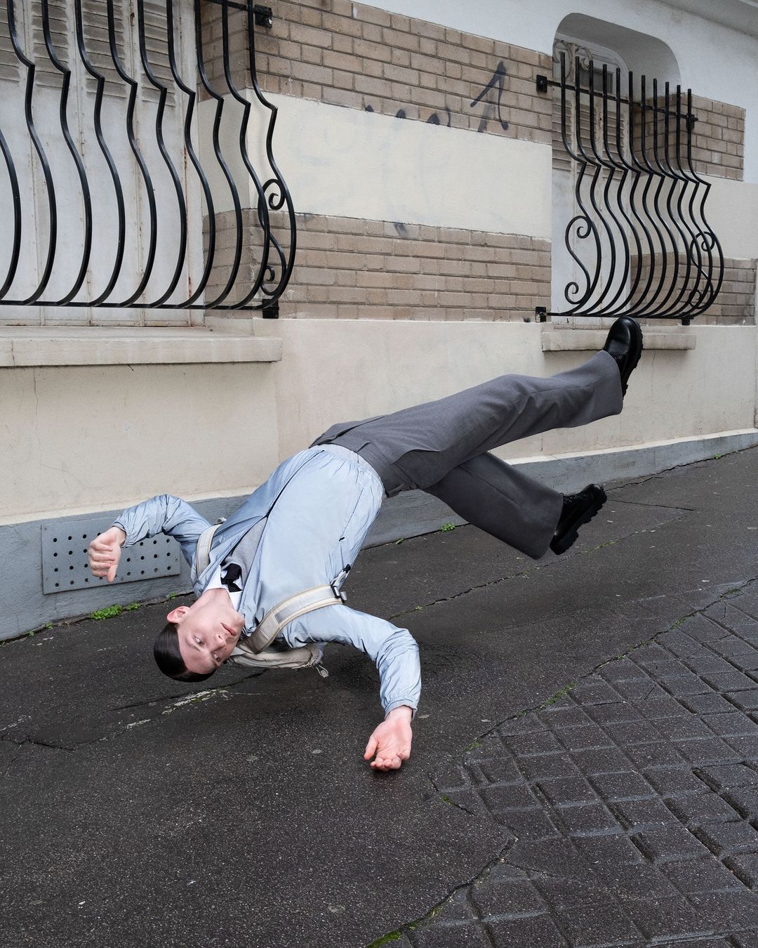 Person in a light blue suit is seemingly suspended above a brick and paved urban street with a textured building facade in the background.