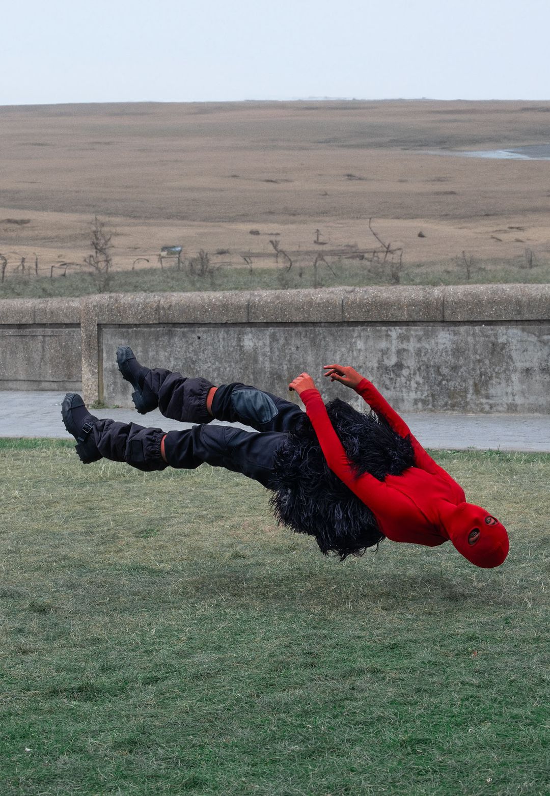 Person in red and black costume appears to float above green grass with a field and overcast sky in the background.