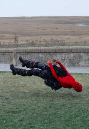 Person in red and black costume appears to float above green grass with a field and overcast sky in the background.