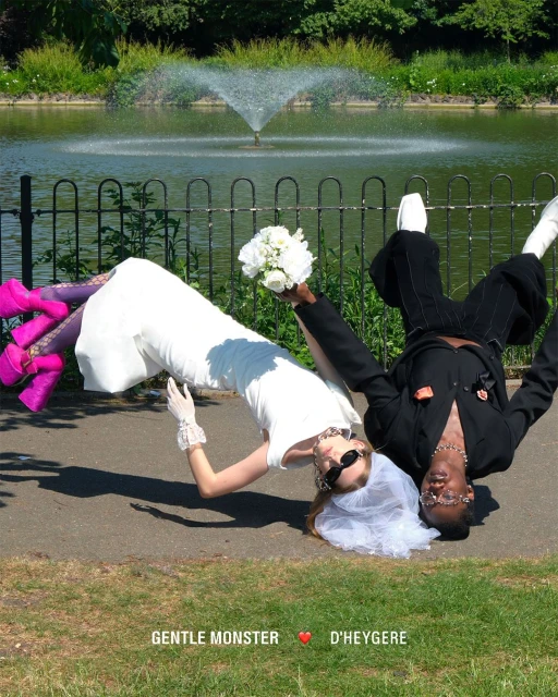 Two people pose upside down in a park near a pond. One is wearing a wedding dress and the other is wearing a black suit.