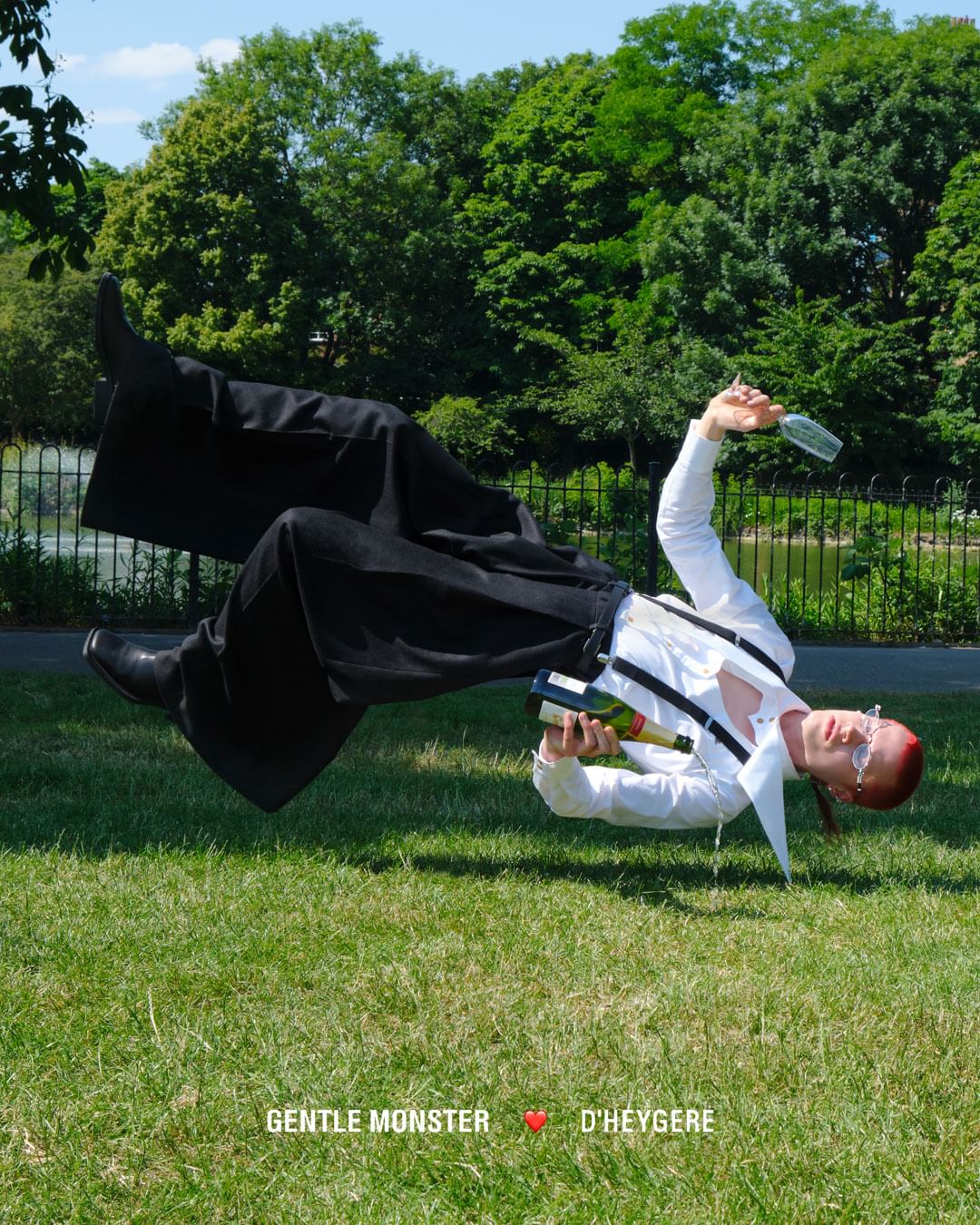 Person in black pants and white shirt with suspenders, holding wine bottle and glass, floating over green grass in outdoor park setting.