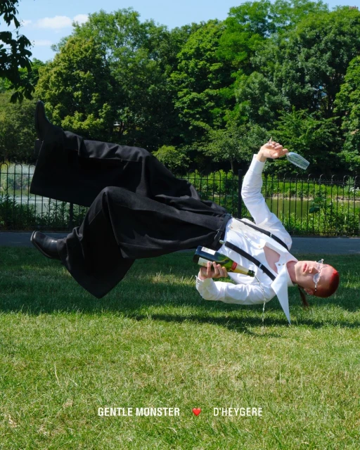 Person in black pants and white shirt with suspenders, holding wine bottle and glass, floating over green grass in outdoor park setting.