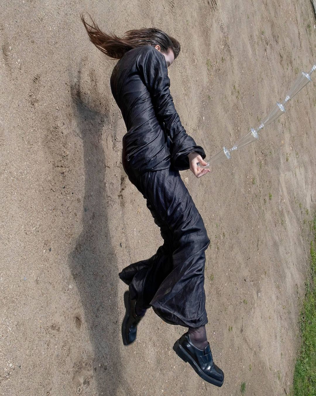 Woman in dark clothing lies on ground outdoors. Long-sleeved top and pants. Hair is long and flowing. Black shoes. Plastic water bottle.