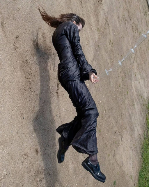 Woman in dark clothing lies on ground outdoors. Long-sleeved top and pants. Hair is long and flowing. Black shoes. Plastic water bottle.