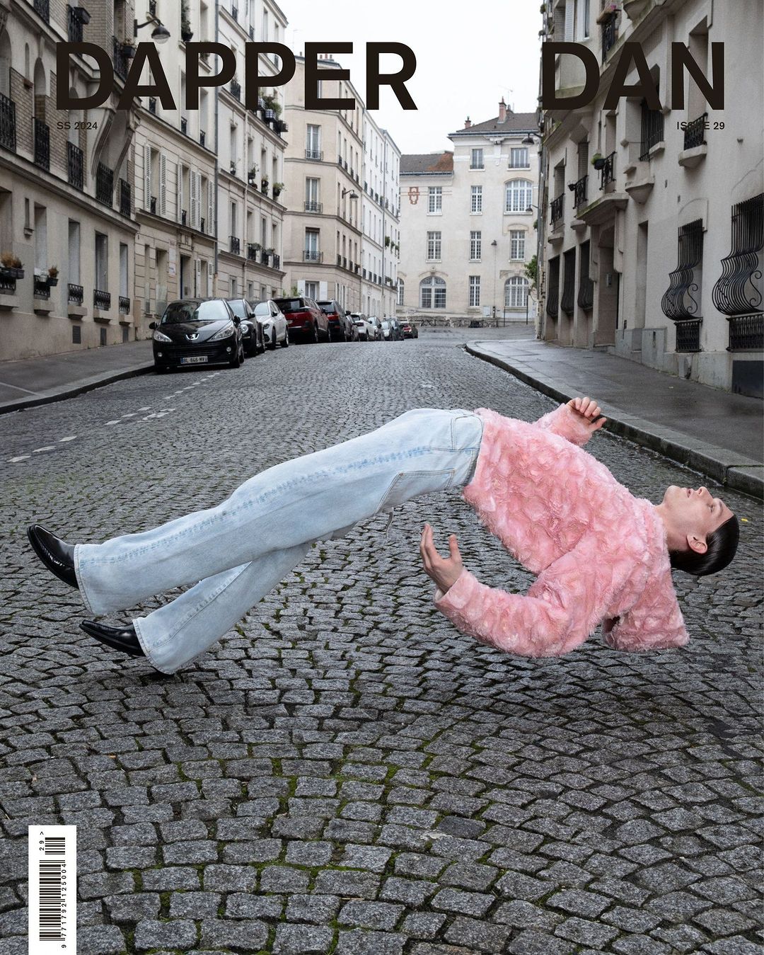 Fashion magazine cover featuring a person in a pink jacket and jeans floating over cobblestones on an urban street with buildings.