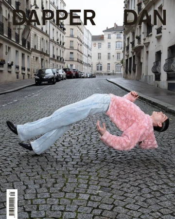 Fashion magazine cover featuring a person in a pink jacket and jeans floating over cobblestones on an urban street with buildings.