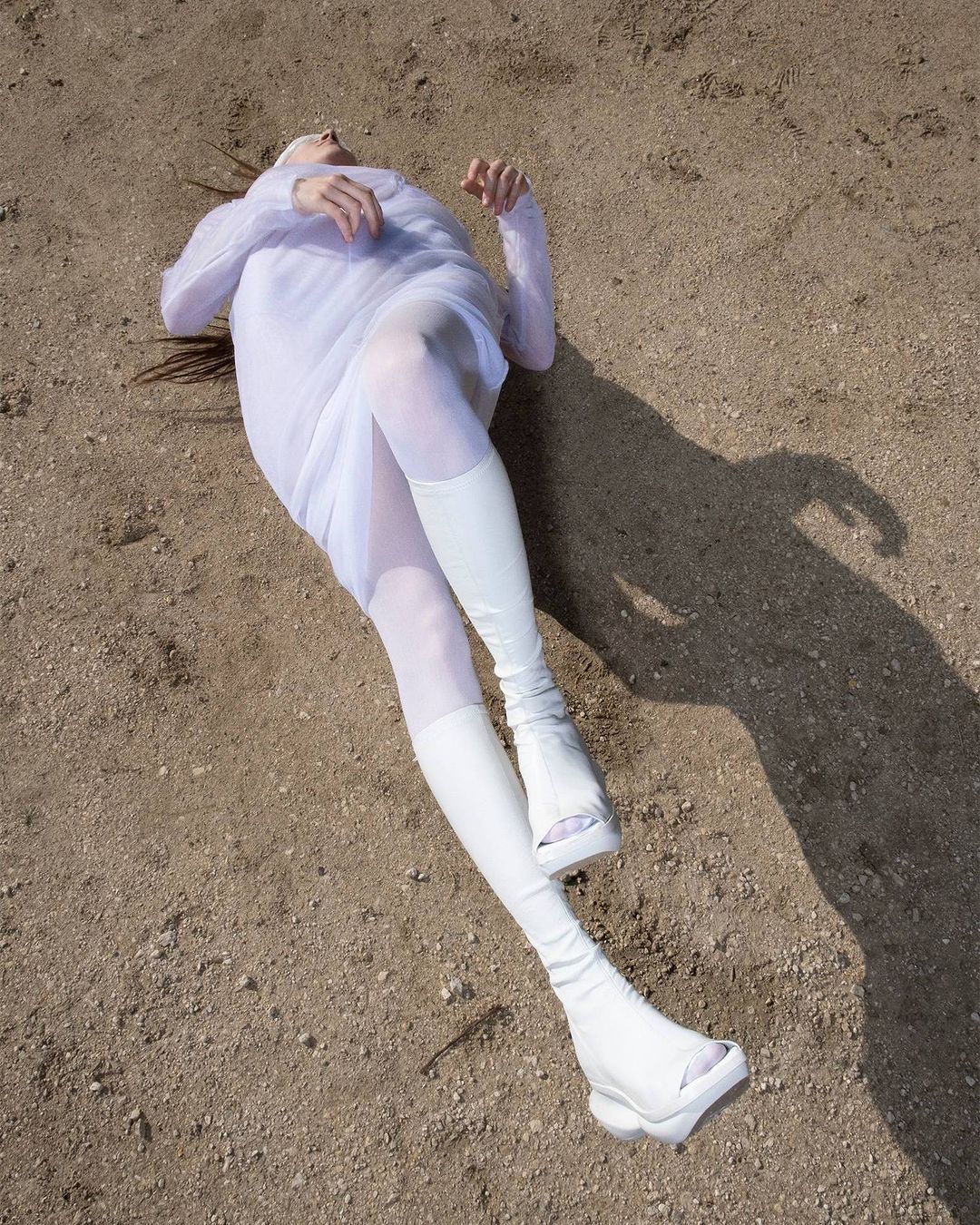 Woman in white lies on sandy ground, wearing dress, boots, and stockings. Full sunlight casts a shadow. Fashion photography.