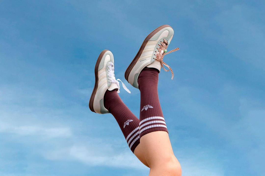 Legs raised against a clear blue sky wearing cream sneakers with patterned laces and burgundy crew socks with white stripes.