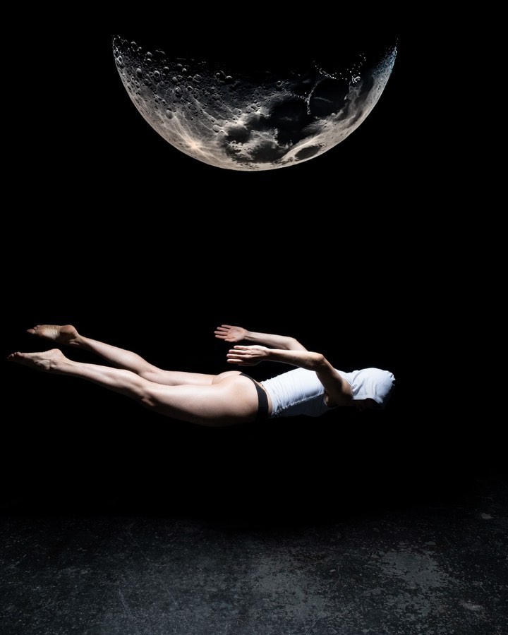 Woman levitates horizontally in a surreal, dark studio shot under a simulated full moon, creating an ethereal, dreamlike aesthetic.