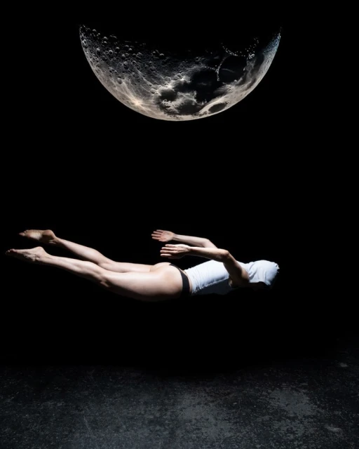 Woman levitates horizontally in a surreal, dark studio shot under a simulated full moon, creating an ethereal, dreamlike aesthetic.