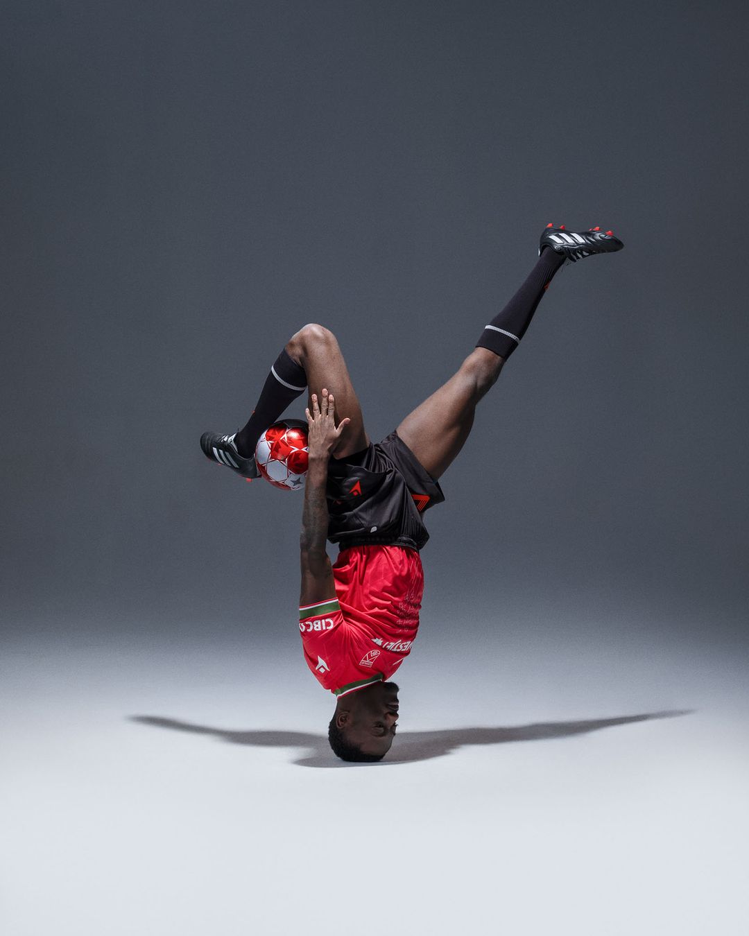 African male soccer player performs handstand balancing a red and white soccer ball on his feet in a studio setting.