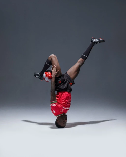 African male soccer player performs handstand balancing a red and white soccer ball on his feet in a studio setting.