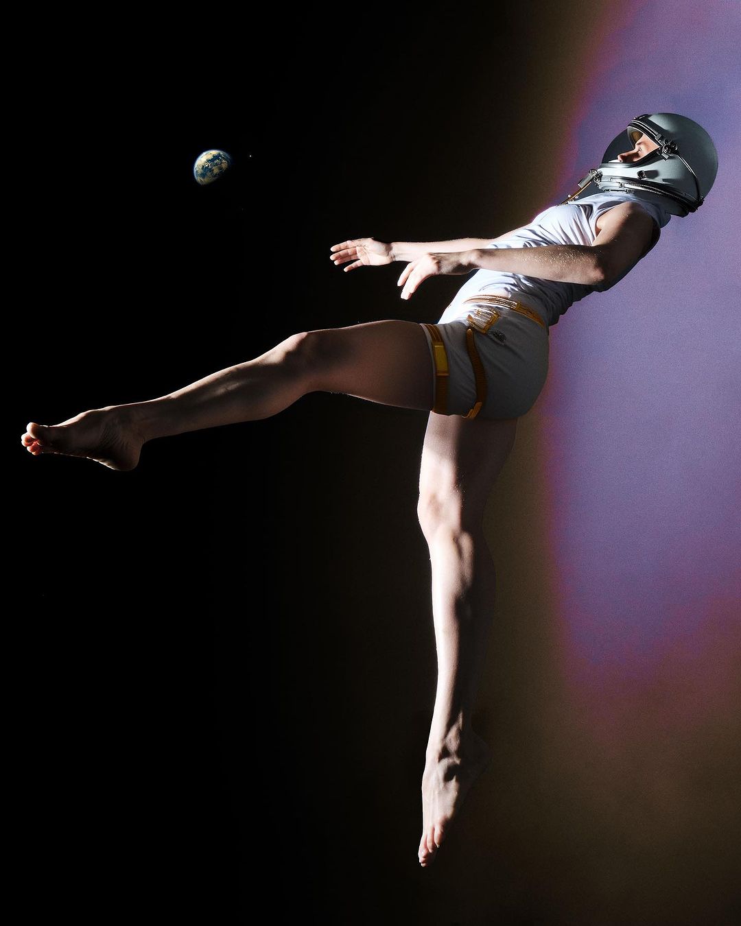 Adult female with a spacesuit helmet performing a kick while suspended in the air against a dark background with a visible Earth.