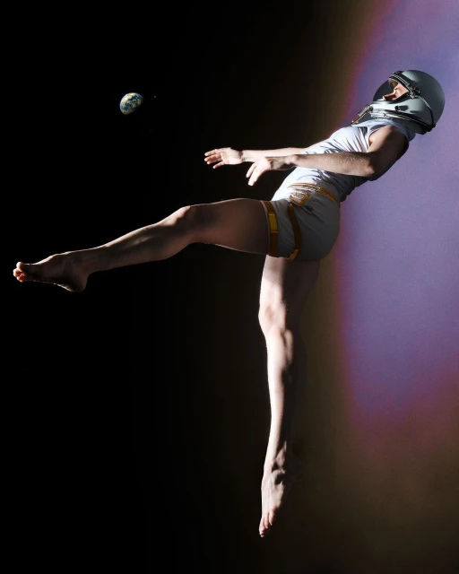 Adult female with a spacesuit helmet performing a kick while suspended in the air against a dark background with a visible Earth.
