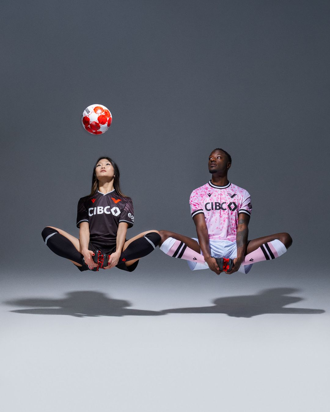 Two soccer players, an Asian woman and a Black man, float cross-legged with a soccer ball above the woman's head in a studio shot.