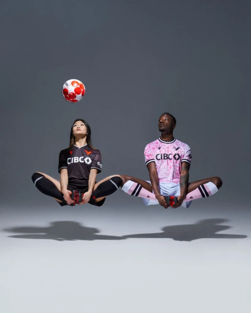 Two soccer players, an Asian woman and a Black man, float cross-legged with a soccer ball above the woman's head in a studio shot.