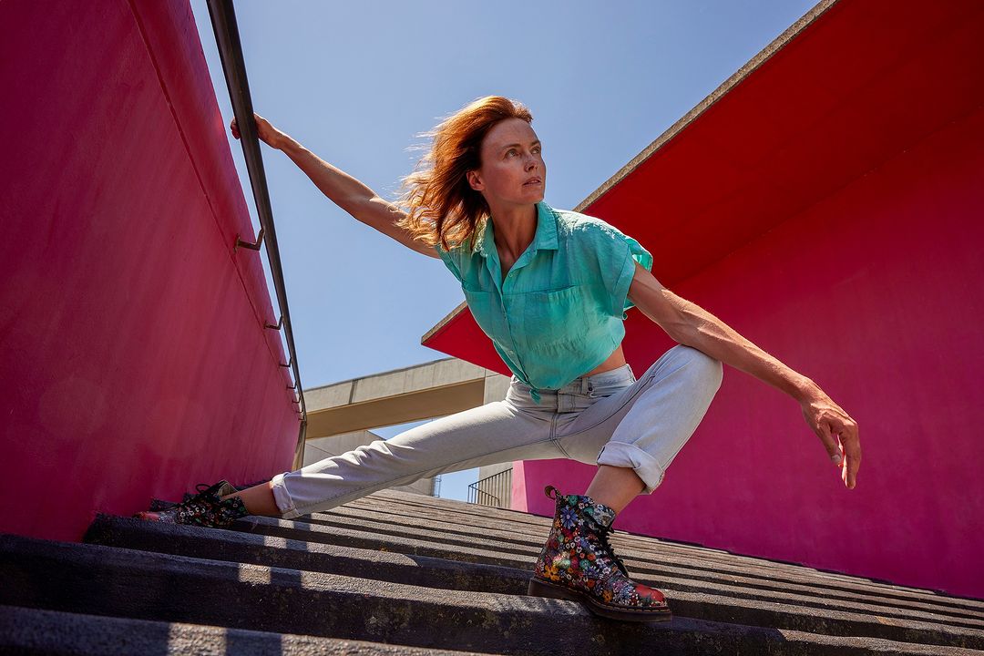 An adult woman with red hair poses on concrete steps in front of a red wall, wearing a teal shirt and flower-print boots in sunlight.