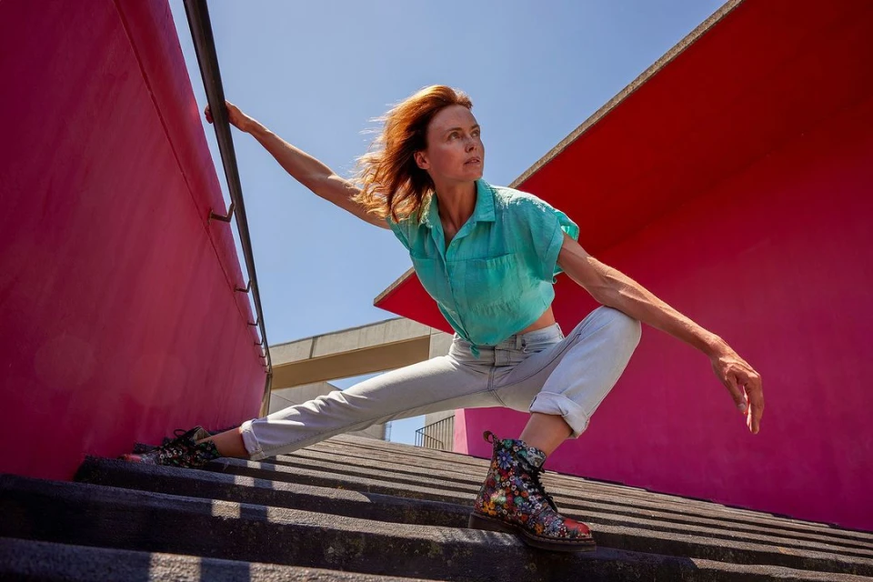 An adult woman with red hair poses on concrete steps in front of a red wall, wearing a teal shirt and flower-print boots in sunlight.