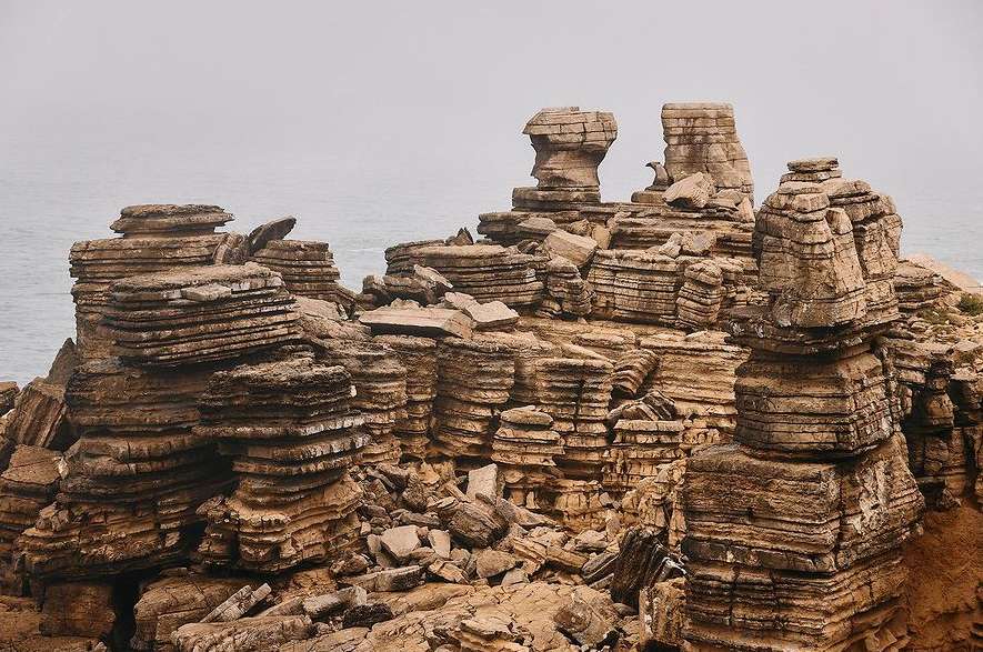 Rocky coastal landscape features layered rock formations near the ocean. The scene is set on an overcast day with a grey sky.