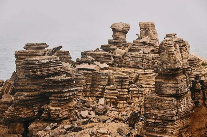 Rocky coastal landscape features layered rock formations near the ocean. The scene is set on an overcast day with a grey sky.