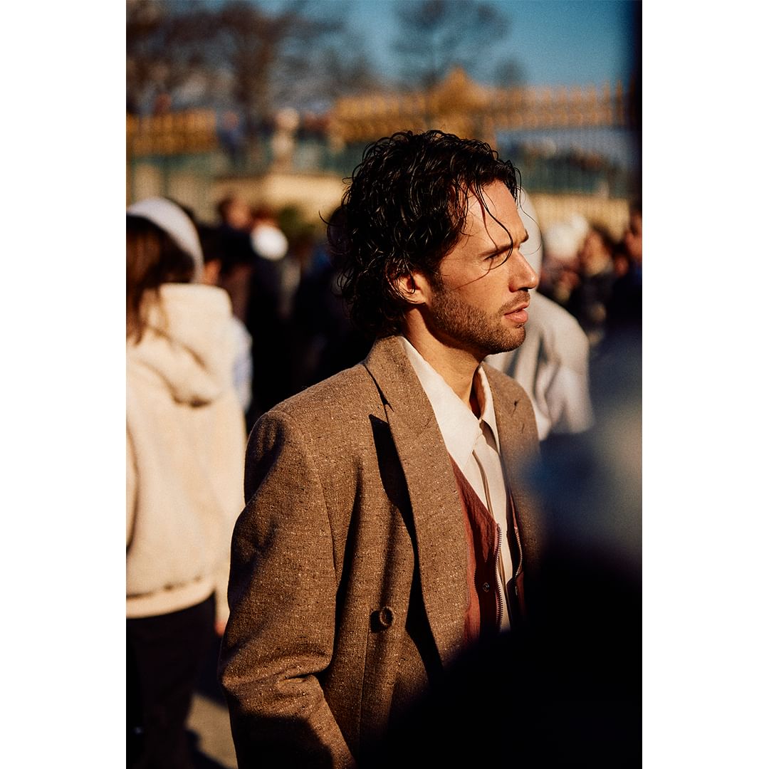 Man in tweed blazer stands in an urban scene, surrounded by a blurred crowd, under natural light. Focus on fashion and people.