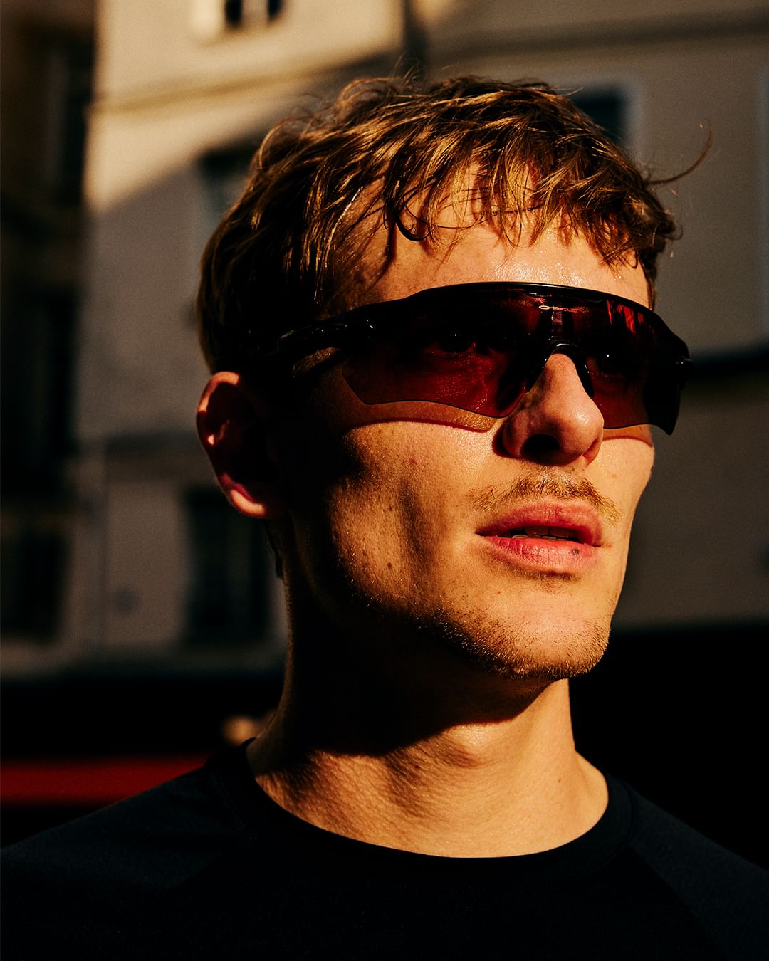 Close-up of a man with stubble wearing dark sunglasses, bathed in warm sunlight against an urban background of apartment buildings.