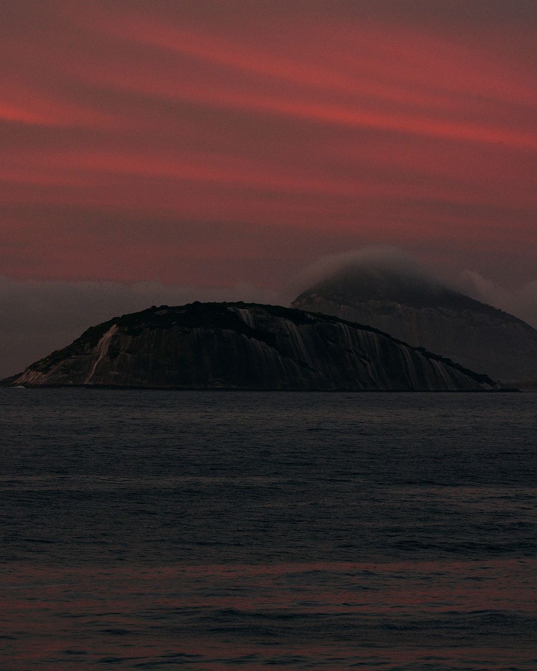 Island in dark water at sunset. Dramatic dusk red and purple sky with clouds. Moody seascape photo of mountain in ocean.