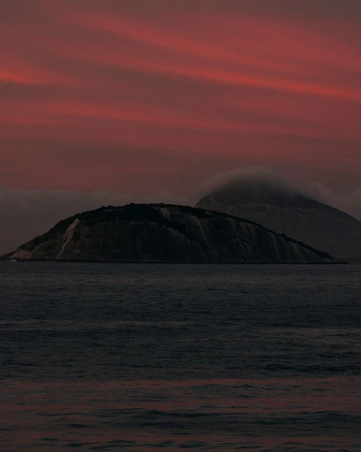 Island in dark water at sunset. Dramatic dusk red and purple sky with clouds. Moody seascape photo of mountain in ocean.