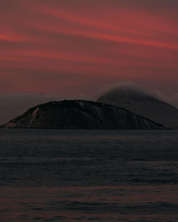 Island in dark water at sunset. Dramatic dusk red and purple sky with clouds. Moody seascape photo of mountain in ocean.