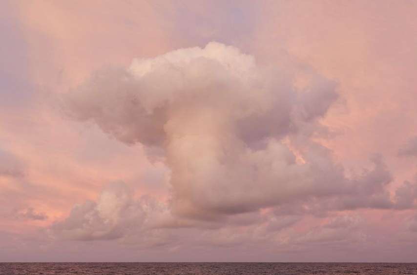 Pink-hued cloudscape featuring an expansive sky with light clouds and a subtle horizon line suggesting a serene seascape below.