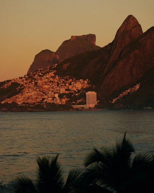 Coastal view featuring Sugarloaf Mountain and Rio de Janeiro architecture during warm sunset lighting, with water in foreground and silhouetted trees.
