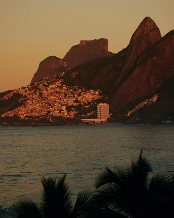 Coastal view featuring Sugarloaf Mountain and Rio de Janeiro architecture during warm sunset lighting, with water in foreground and silhouetted trees.