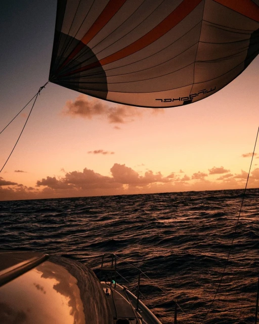 Sailboat on the ocean at sunset, showing a large sail, ocean waves, and colorful sunset sky, creating a sense of adventure and tranquility.