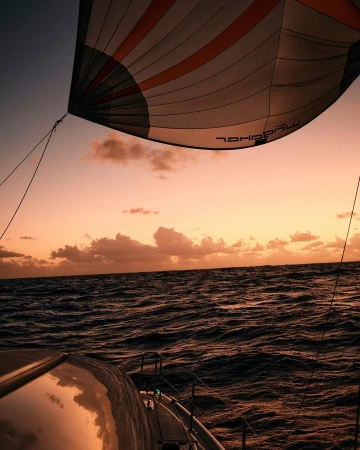Sailboat on the ocean at sunset, showing a large sail, ocean waves, and colorful sunset sky, creating a sense of adventure and tranquility.