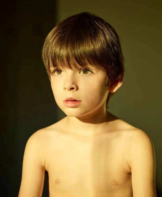 Boy with brown hair and a thoughtful expression, lit by golden sunlight, posing indoors against a dark olive background in a medium shot.