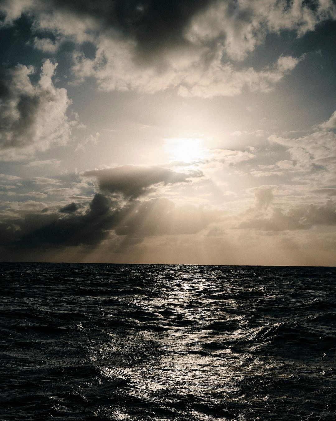 Dramatic ocean scene with dark, choppy water reflecting sunlight piercing through a cloudy sky, creating a contrast of light and shadow.