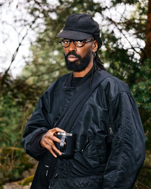 Man with beard and dreadlocks wearing a baseball cap and glasses holds a mug outdoors surrounded by pine trees in a medium shot.