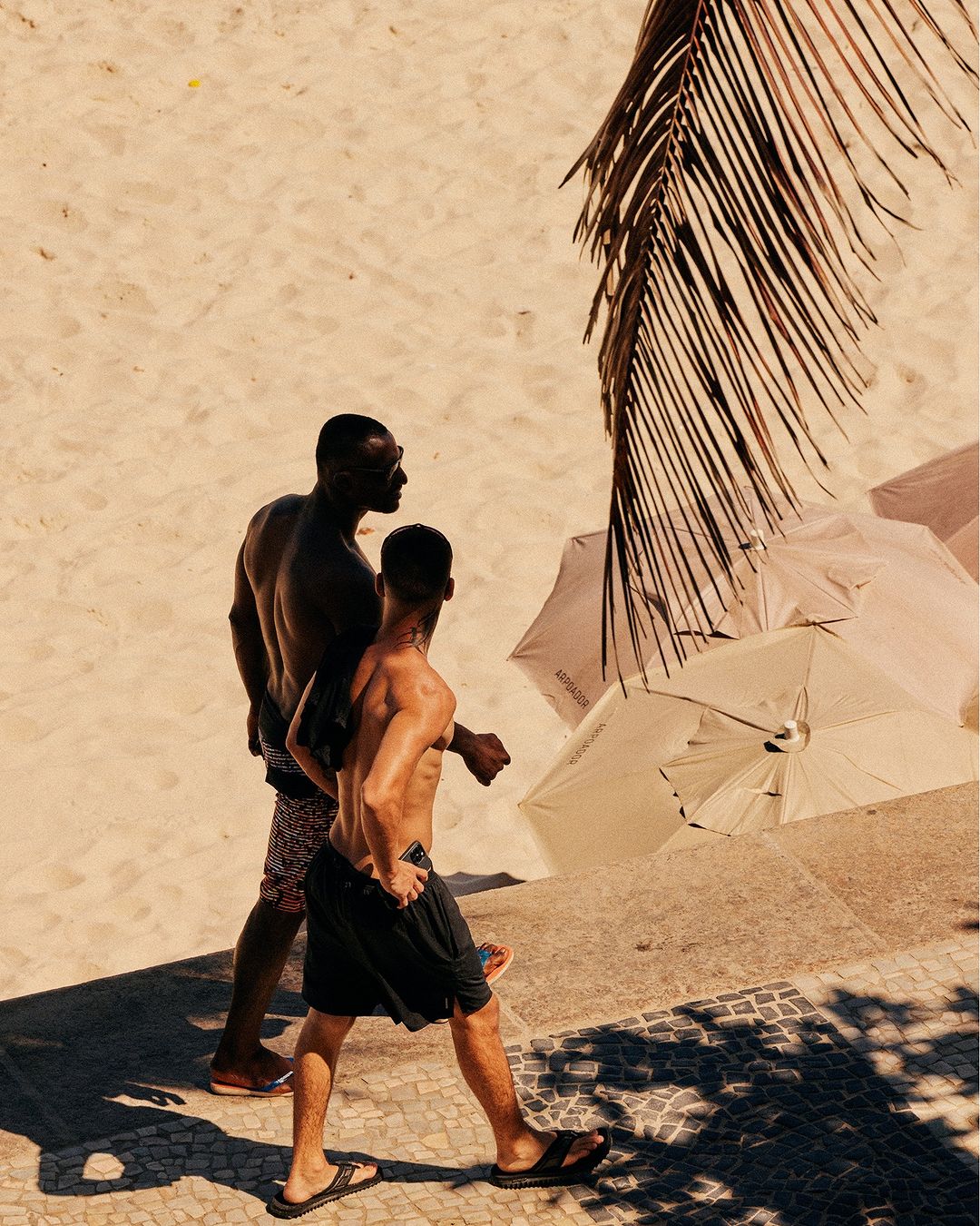 Two shirtless men walking on a tiled promenade along a sandy beach, with a palm frond and umbrellas in the background.