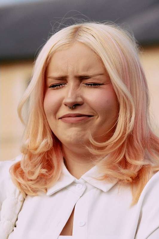 Close-up portrait of an adult female with a slight grimace expression, blonde and peach wavy hair, wearing a white collared shirt.